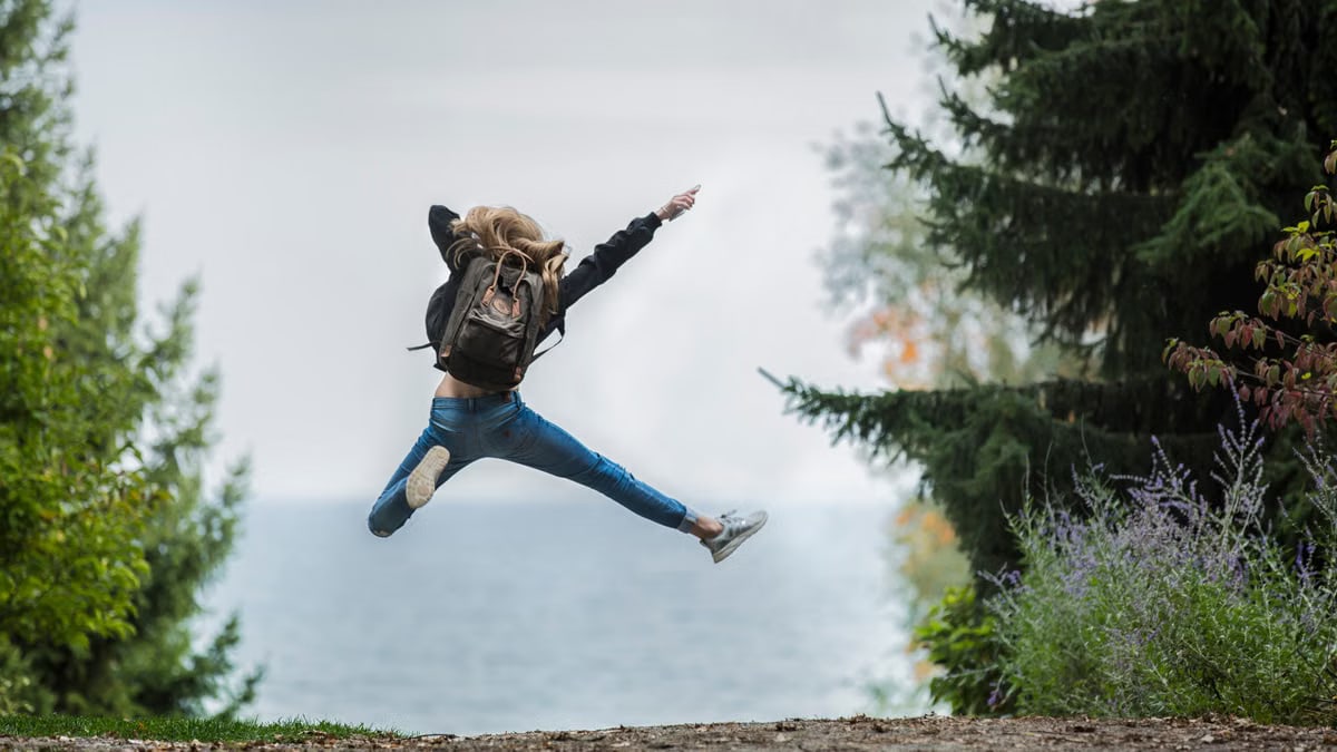 Energetic woman leaping in outdoor forest setting with lake view, showcasing freedom and joy.